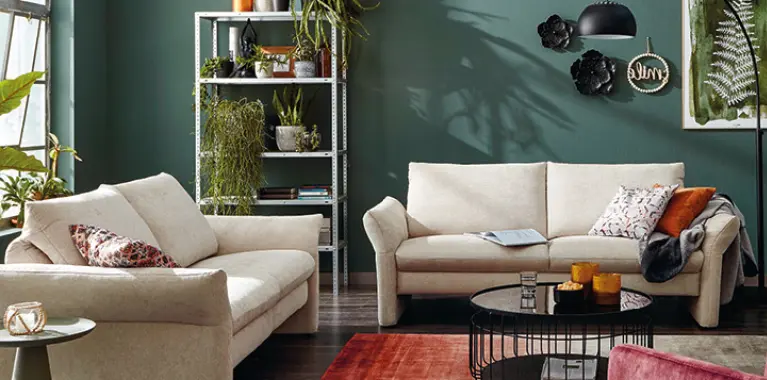 Zwei cremefarbene Sofas vor gruener Wand, Wanddeko links, schwarzer runder Couchtisch, Regal mit Pflanzen rechts, gemusterter Teppich